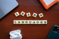 Klocki z literami układające napis „LEARN LANGUAGES” na drewnianym biurku. Wokół widoczne są laptop, smartfon, słuchawki bezprzewodowe oraz dysk zewnętrzny.