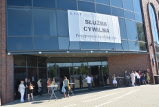 głowne wejście do budynku EXPO XXI, nad wejściem duży napis postępowanie kwalifikacyjne w służbie cywilnej, przed wejściem stoją uczestnicy i rozmawiają między sobą przy stolikach