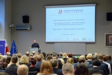 Uczestnicy seminarium w auli KSAP, Prorektor ds. studiów Wyższej Szkoły Policji w Szczytnie przemawia przy mównicy, w tle na dużym ekranie wyświetlona prezentacja z tytułem i logo seminarium