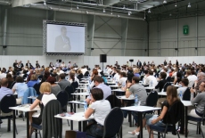 Sala egzaminacyjna, kandydaci siedzą przy stolikach i czekają na rozpoczęcie egzaminu.